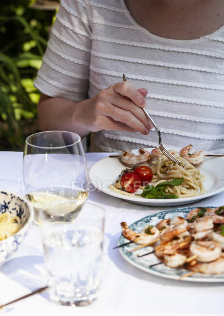 Femme tourbillonnant pâtes cacio e pepe sur une assiette avec brochettes de crevettes, tomates et vin blanc à une table extérieure — Photo de stock