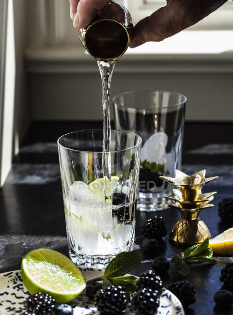 Hand pouring shot glass of vodka into cocktail glass with blackberries, lime and mint — Stock Photo