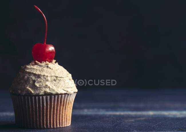 A cupcake with peanut frosting and a cocktail cherry in front of a dark background — Stock Photo