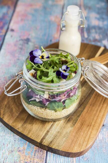 Un'insalata di quinoa con agnelli lattuga, radicchio, rucola, crostini, formaggio di capra e viola cornuto in un barattolo di vetro su una tavola di legno, con condimento in una bottiglia di vetro — Foto stock