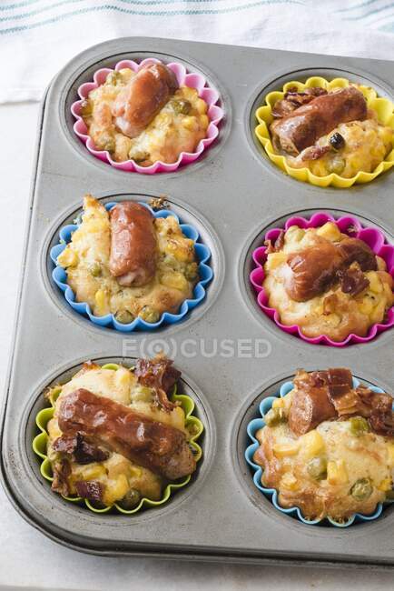A metal muffin tray with savoury Frankfurter sausage muffins in colourful silicone cases. Crispy bacon, corn and green peas — Stock Photo