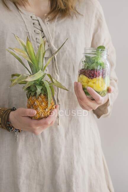 A woman in a linen dress holding a jar of fruit salad with mango, pomegranate seeds, lettuce and a fresh baby pineapple — Stock Photo