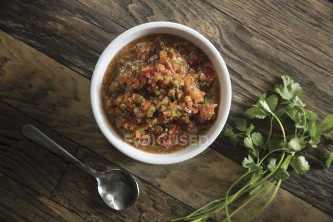 Gazpacho de tomate en un tazón blanco sobre una superficie de madera junto a un cilantro - foto de stock