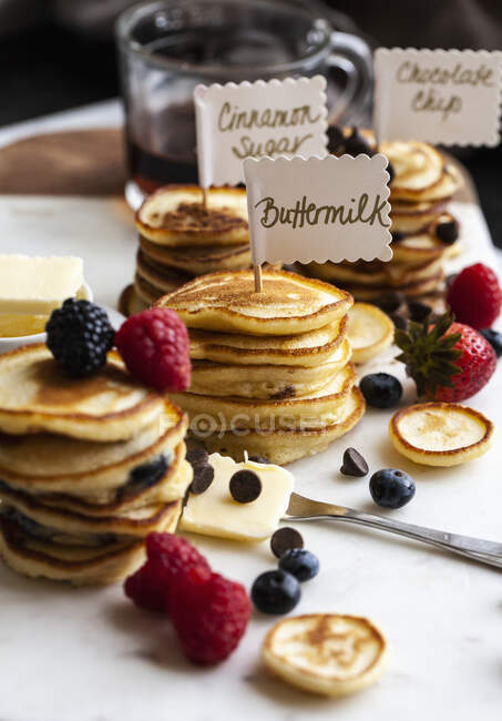 Pile di mini pancake con cartelli, con gocce di frutta e cioccolato, sciroppo e burro, su una tavola di marmo — Foto stock