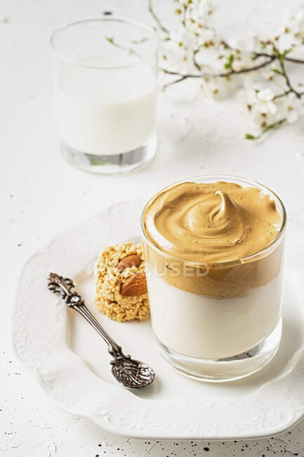 Dalgona coffee in a transparent glass with a spoon and cookies on a white plate with a bunch of blooming apple tree — Stock Photo