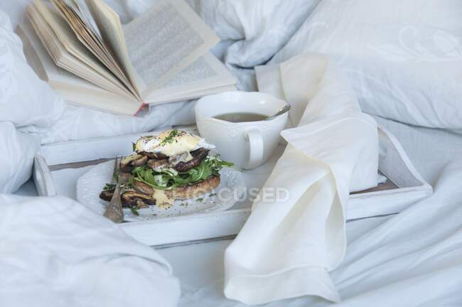 Ovos Benedict com cogumelos na bandeja do café da manhã na cama com xícara de chá e livro — Fotografia de Stock