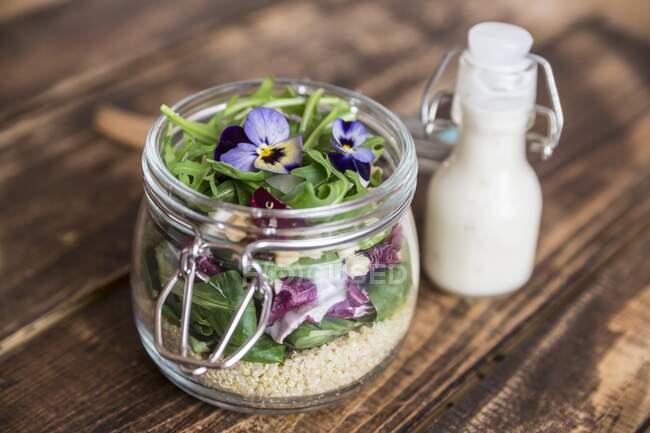 A quinoa salad with lambs lettuce, radicchio, rocket, croutons, goat's cheese and horned violets in a glass jar, with dressing in a glass bottle — Stock Photo