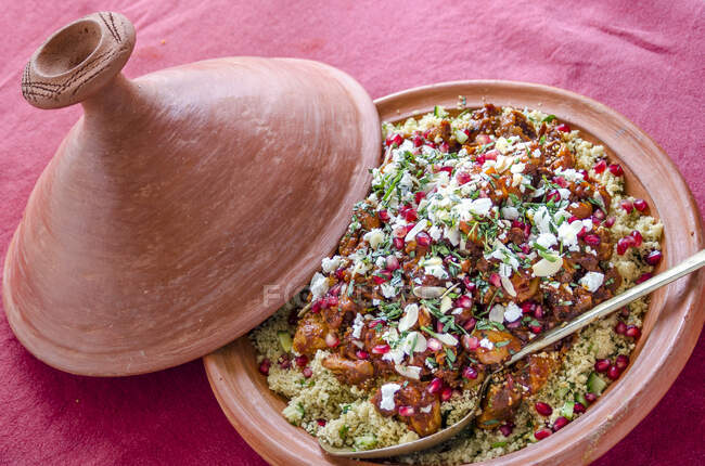 Chicken cous cous tagine with pomegranate seeds, feta cheese and herbs in a terracotta tagine with a serving spoon on a red tablecloth — Stock Photo