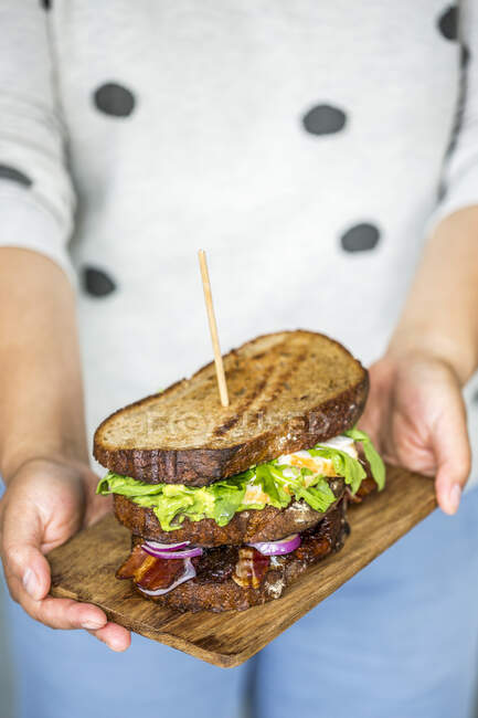 Person holding wooden board with grilled sandwich with vegetables, egg and bacon — Stock Photo