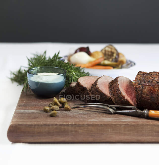 Rolled roast beef with horseradish sauce, rosemary and capers, roast vegetables in the background — Stock Photo
