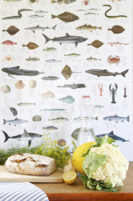 Du pain, du chou-fleur, de l'aneth, du melon, du citron et une carafe d'eau devant du papier peint de poisson — Photo de stock