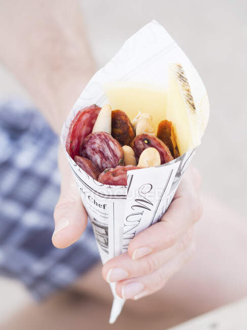 Hand holding paper cone with selection of Manchego cheese and different salami types — Stock Photo