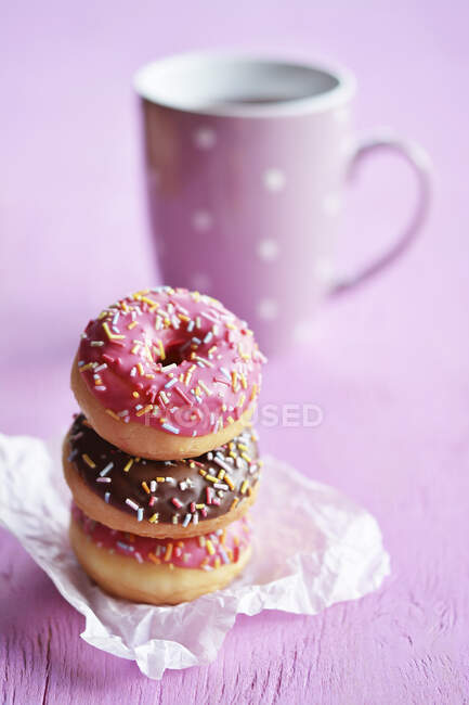 A stack of three mini doughnuts with icing and sugar strands in front of a teacup — Stock Photo