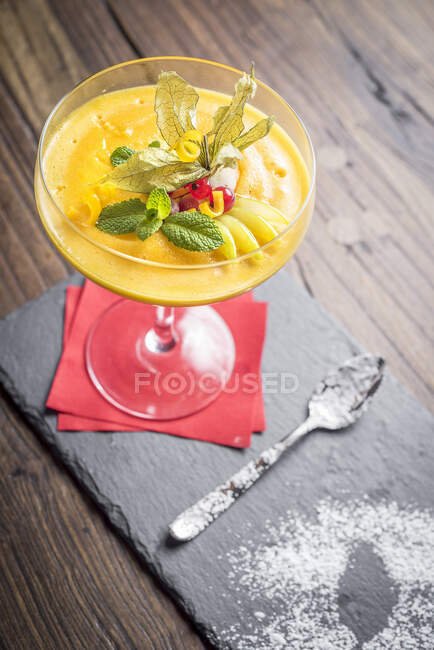 Sorbete de mango decorado con cáscara de cítricos, menta, grosella roja en un vaso con una servilleta roja y una cuchara sobre una pizarra y fondo de madera - foto de stock