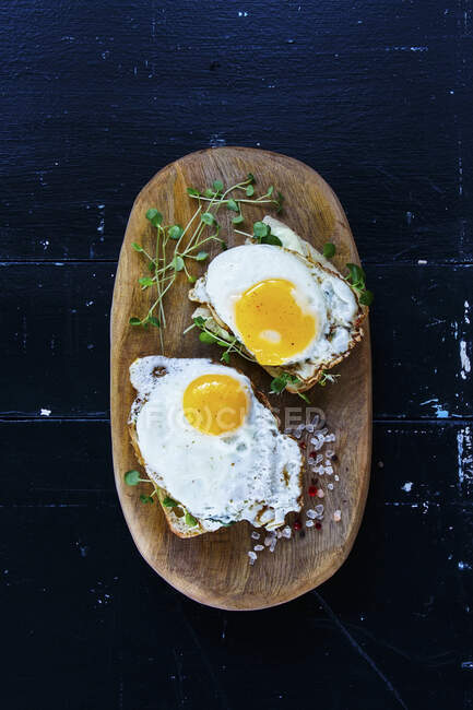 Closen up of breakfast toast with fried eggs and sprouts on wooden board over black grunge background — Stock Photo
