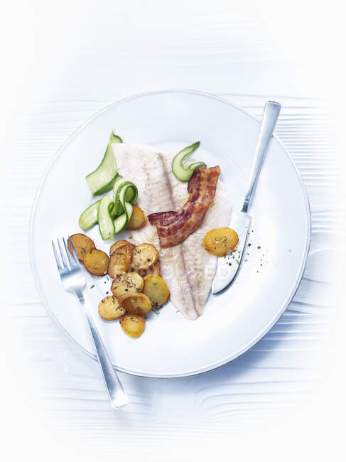 Bacon and fish fillet with roast potatoes and cucumber on plate with cutlery — Stock Photo