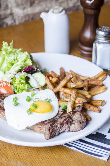 Beef rump steak topped with fried egg and chives herbs served with golden chips and fresh salad on a white plate on a wooden table — Stock Photo