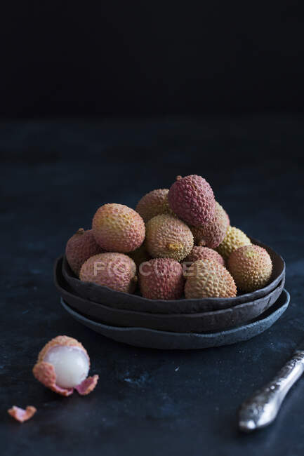 Lychees in dark bowl and peeled on table with knife — Stock Photo