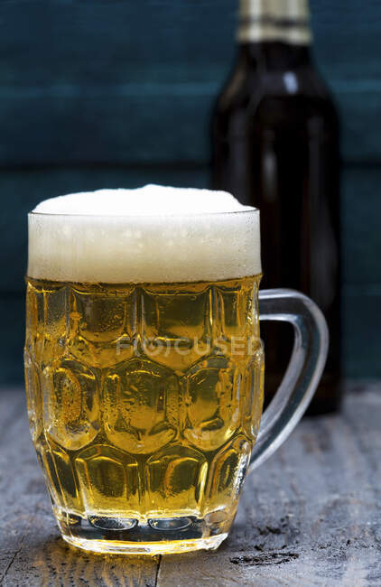 Birra leggera in vetro con schiuma e bottiglia sullo sfondo — Foto stock