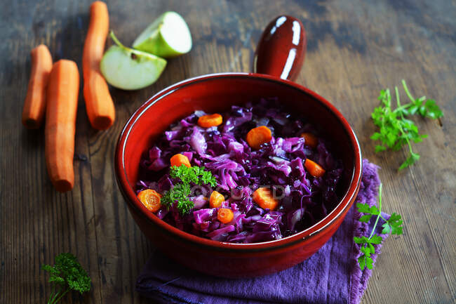 Red cabbage with carrots and apple in a ceramic pot with fresh ingredients in the background — Stock Photo