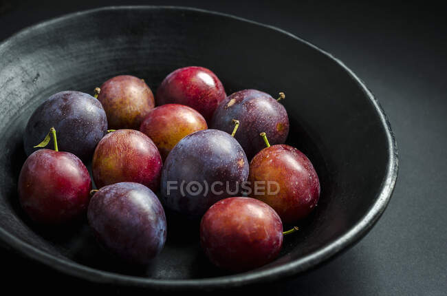 Ciotola di prugne viola e rosse fresche e prugne in una ciotola di frutta nera su uno sfondo nero — Foto stock
