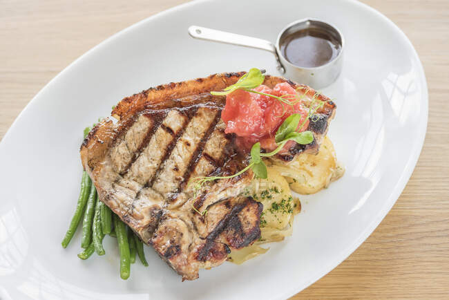 Steak grillé avec de longs haricots verts, pommes de terre et tomates grillées avec sauce steak sur une assiette blanche et un fond en bois clair — Photo de stock