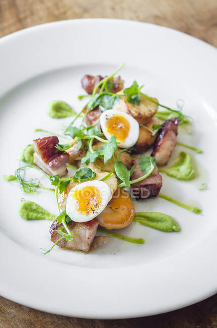 Pan fried bacon and scallops with quail eggs and a pea puree with pea shoots on a white plate and wooden table — Stock Photo