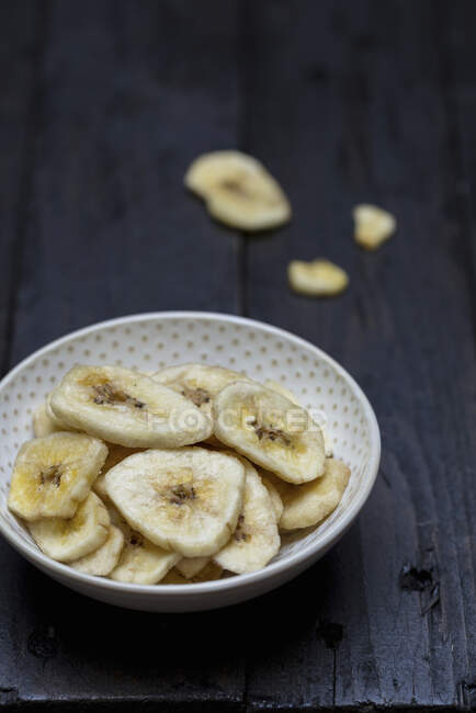 Banana chips in bowl and on black wooden table — Stock Photo