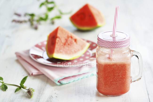 Jug with watermelon smoothie and straw in front of plate with melon pieces and fresh mint — Stock Photo
