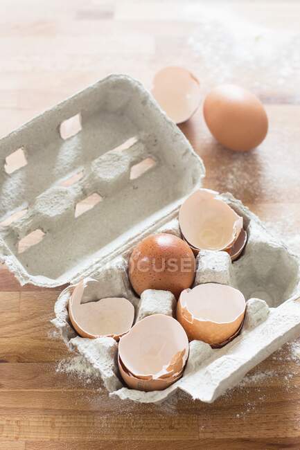 Бумажная коробка с оболочками и яйцами на деревянном столе с мукой — стоковое фото