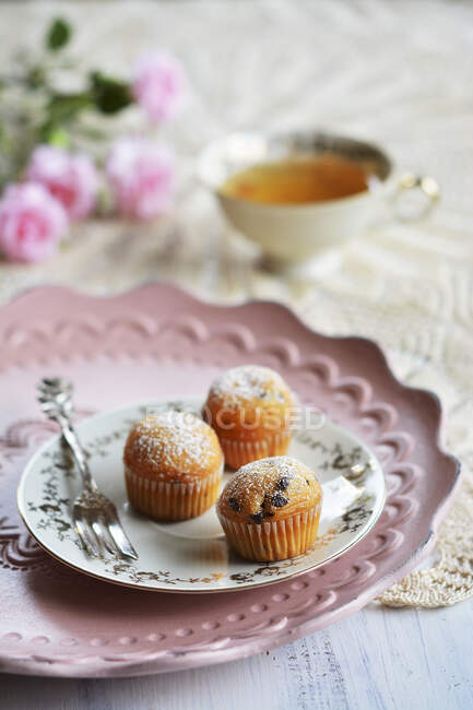 Las magdalenas pequeñas en el plato con el tenedor, la taza de té y las flores - foto de stock