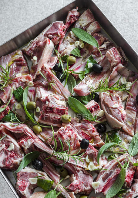 Pieces of raw lamb with garlic, olives, sage, rosemary and bay leaves on a baking tray — Stock Photo