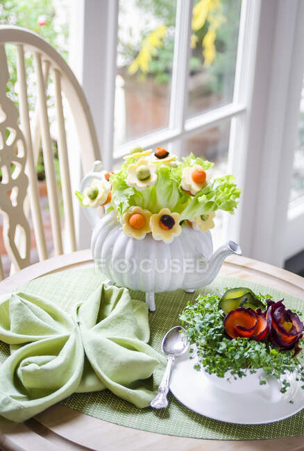 Ramo de hojas de ensalada y flores hechas con queso, zanahorias y aguacate en una tetera, berro y zanahorias en una taza de té sobre una manta verde - foto de stock