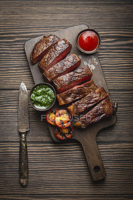 Grilled or fried and sliced marbled meat steak with fork, tomatoes as a side dish and different sauces on wooden cutting board, top view, close-up — Stock Photo
