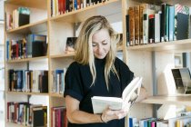 Женщина читает книгу, опираясь на книжную полку — стоковое фото
