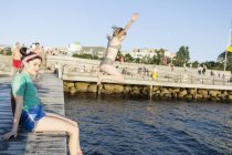 Femme assise sur la jetée avec un ami sautant — Photo de stock