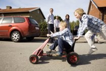Мальчик толкает брата на четырехколесный цикл с семьей на заднем плане — стоковое фото