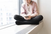 Девушка сидит на подоконнике со смартфоном — стоковое фото