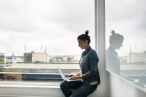 Вид сбоку на женщину, использующую ноутбук на подоконнике в офисе — стоковое фото