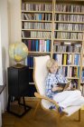 Niño leyendo libro en silla de salón en frente de estanterías en el interior del hogar - foto de stock