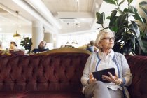 Старшая женщина с цифровым планшетом сидит на диване и смотрит в сторону — стоковое фото