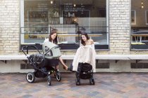 Dos madres jóvenes con cochecitos de bebé sentadas frente a la cafetería - foto de stock