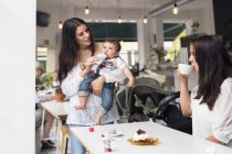 Mother holding baby son (6-11 months) and talking with friend in cafe — Stock Photo
