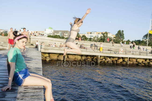 Жінка сидить на пірсі з другом стрибає — стокове фото
