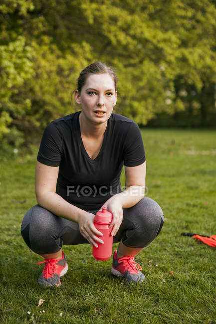 Mulher segurando garrafa de água enquanto se agacha no campo — Fotografia de Stock