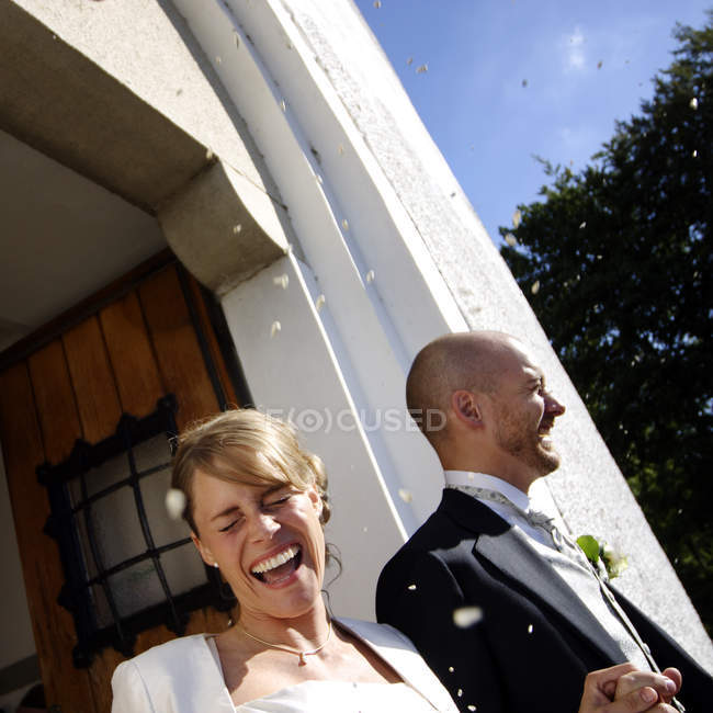 Recém-casados alegres andando fora da igreja sob chuva de confetes — Fotografia de Stock