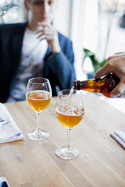 Vista recortada de la mano de la persona vertiendo cerveza en vasos con el hombre desenfocado sentado en el restaurante - foto de stock