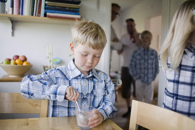 Niño revolviendo cuchara en vidrio en la mesa en casa con la familia en el fondo - foto de stock