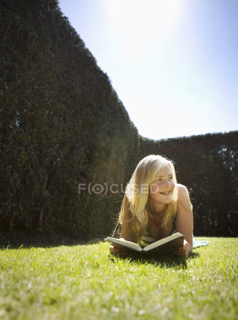 Visão frontal da menina adolescente lendo livro na grama no parque — Fotografia de Stock