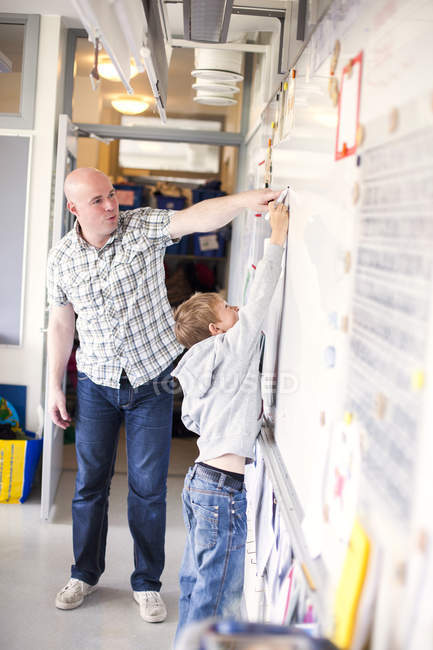 Homme enseignant aider garçon dans l'écriture sur tableau blanc à la salle de classe — Photo de stock